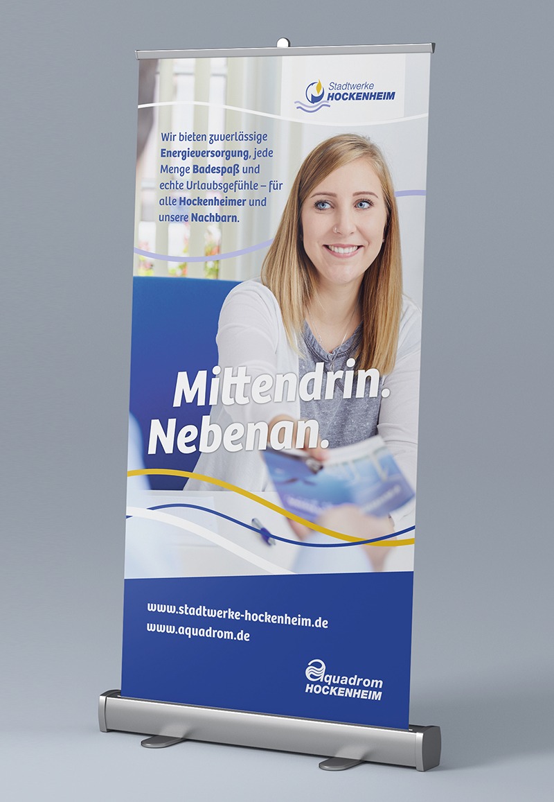 Auf einem Roll-Up-Banner der Stadtwerke Hockenheim ist der prominente Slogan „Mittendrin. Nebenan.“ und eine lächelnde Frau zu sehen, die eine Broschüre entgegennimmt. Der Claim ist Dreh- und Angelpunkt der Marketingstrategie.