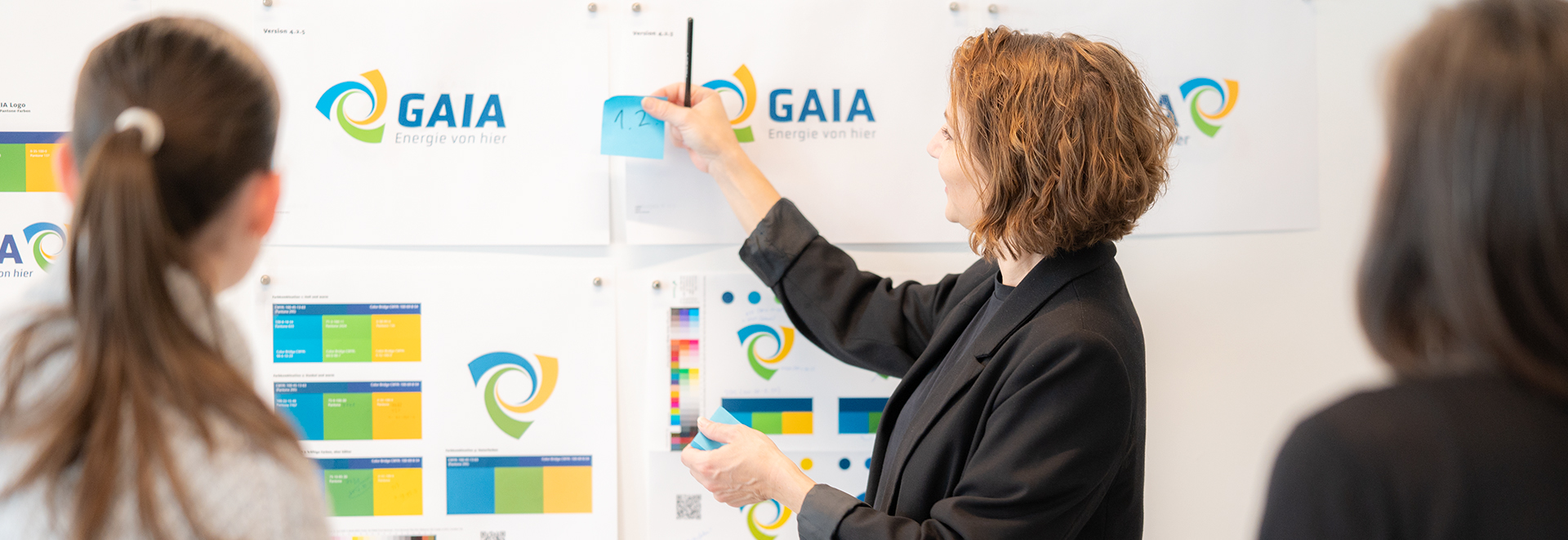 Creative Director präsentiert in Workshop Logoentwürfe an einer Wand mit Farbmustern, während zwei Kund:innen aufmerksam zusehen.